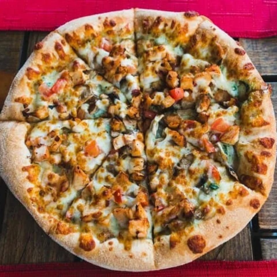 A whole, sliced chicken pizza with melted cheese, tomatoes, and mushrooms on a wooden table.