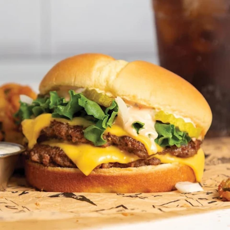 Double cheeseburger with lettuce, pickles, and sauce on a bun, with onion rings and a drink.